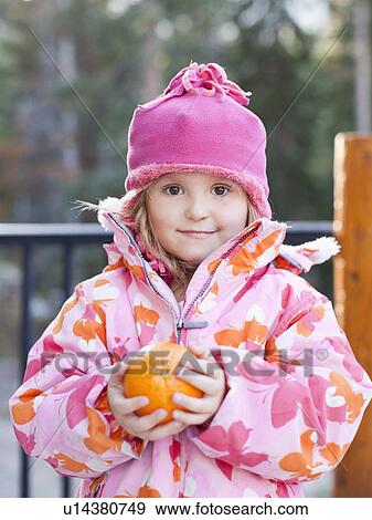Stock Photo - Girl (4-5) wearing warm clothing holding orange outdoors. Fotosearch