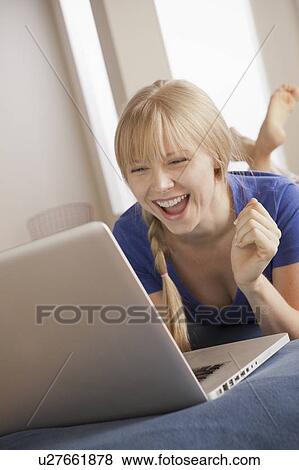 Stock Photo - Laughing young woman lying on bed using laptop. Fotosearch
