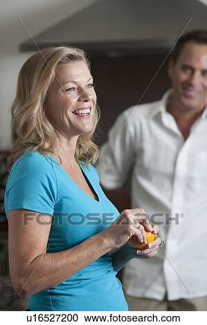 Stock Image - Portrait of mature woman eating orange, man in background. Fotosearch