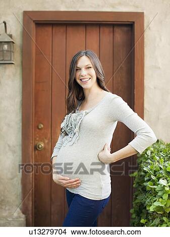 Picture - Portrait of pregnant mid adult woman standing in front of entrance door. Fotosearch