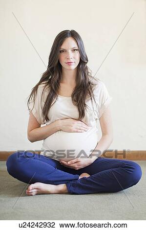 Stock Image - Portrait of pregnant mid adult woman sitting with crossed legs touching her abdomen. Fotosearch