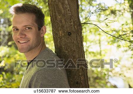 Stock Photo - Portrait of young man leaning against tree trunk. Fotosearch