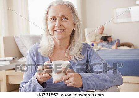 Stock Image - Senior woman holding coffee cup with husband reading in background. Fotosearch