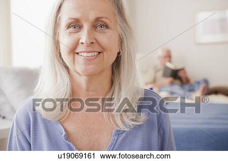 Senior woman smiling with husband reading in background View Large Photo Image Stock Image - Senior woman smiling with husband reading in background. Fotosearch