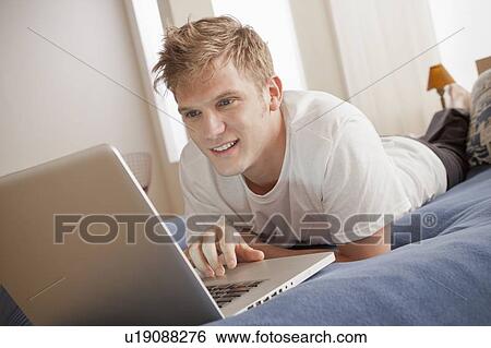Stock Photograph - Smiling man lying on bed using laptop. Fotosearch