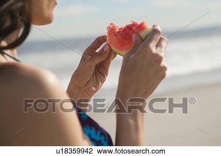 Stock Image - Woman eating watermelon at beach. Fotosearch