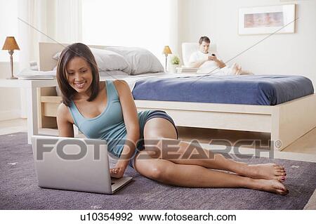 Stock Image - Woman using laptop with husband on lounger in background. Fotosearch