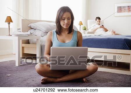 Stock Image - Woman using laptop with husband on lounger in background. Fotosearch