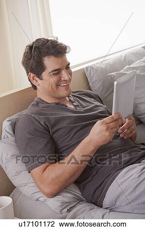 Stock Image - Young man laying on bed and using digital tablet. Fotosearch