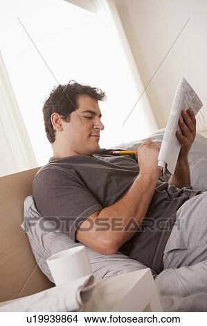 Picture - Young man laying on bed filling crossword. Fotosearch