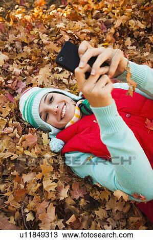 Stock Image - Young woman lying on autumn leaves and using . Fotosearch