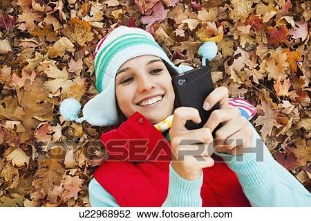 Stock Image - Young woman lying on autumn leaves and using . Fotosearch