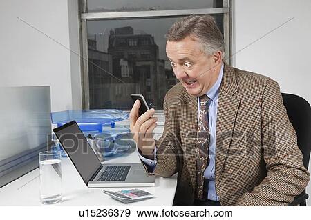 Angry middle-aged businessman looking at cell phone in office View Large Photo Image Stock Photo - Angry middle-aged businessman looking at cell phone in office. Fotosearch