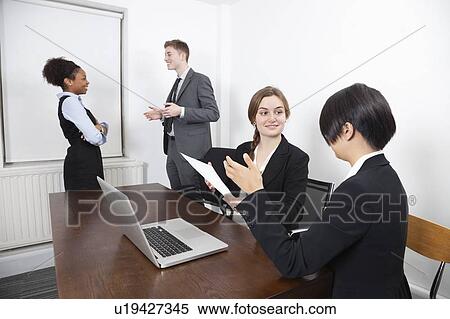 Multiethnic females with document and laptop while colleagues conversing in background View Large Photo Image Stock Photography - Multiethnic females with document and laptop while colleagues conversing in background. Fotosearch