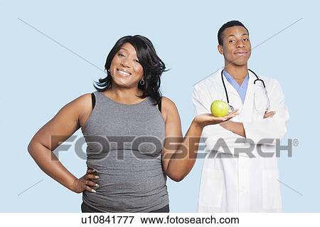 Stock Photo - Overweight mixed race woman holding green apple with doctor over blue background. Fotosearch