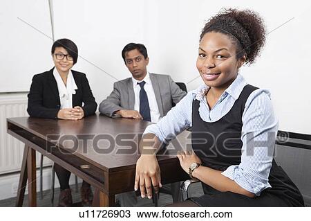 Stock Image - Portrait of African American female with colleagues in background at desk in office. Fotosearch