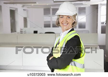 Stock Image - Portrait of smiling senior woman in reflector vest and hard hat at office. Fotosearch