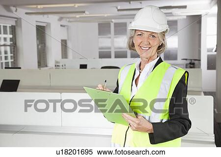 Stock Photography - Portrait of smiling senior woman in reflector vest and hard hat holding clipboard at office. Fotosearch