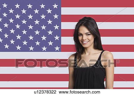 Stock Image - Portrait of young woman smiling against American flag. Fotosearch