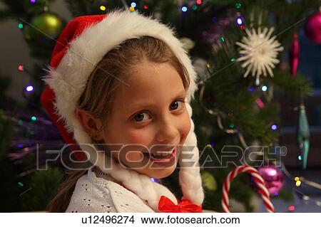 Picture - Girl wearing Santa hat by Christmas tree. Fotosearch