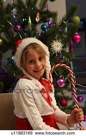 Stock Photo - Girl wearing Santa hat by Christmas tree. Fotosearch