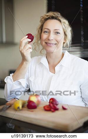 Stock Image - Woman eating fruit in kitchen. Fotosearch