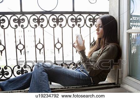 Stock Image - Woman using cell phone in windowsill. Fotosearch