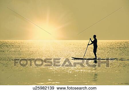 Man standing on paddle board at sunset View Large Photo Image Stock Photo - Man standing on paddle board at sunset. Fotosearch