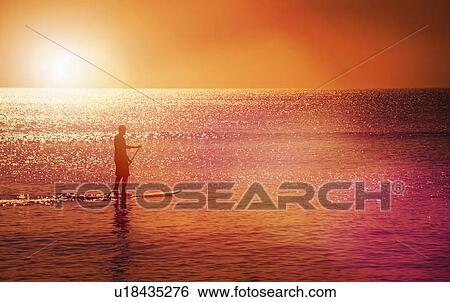 Stock Photograph - Man standing on paddle board at sunset. Fotosearch