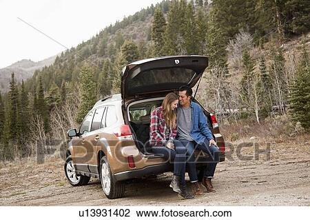 Stock Image - Portrait of young couple sitting in car trunk in non-urban scene. Fotosearch