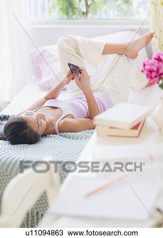 Stock Image - View of Teenage girl (16-17 years) using mobile phone next to desk with books. Fotosearch
