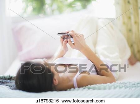 Stock Photograph - View of Teenage girl ( 16-17 years) using mobile phone lying on bed. Fotosearch