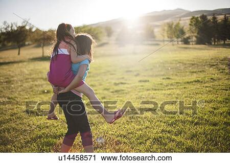 Stock Image - Girl carrying friend piggyback in field. Fotosearch