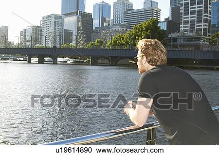 Stock Image - Man overlooking urban river on bridge. Fotosearch