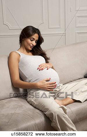 Stock Image - Pregnant woman holding belly on sofa. Fotosearch