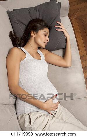 Stock Photo - Pregnant woman holding belly on sofa. Fotosearch