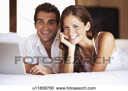 Stock Image - Couple using laptop while lying in bed. Fotosearch