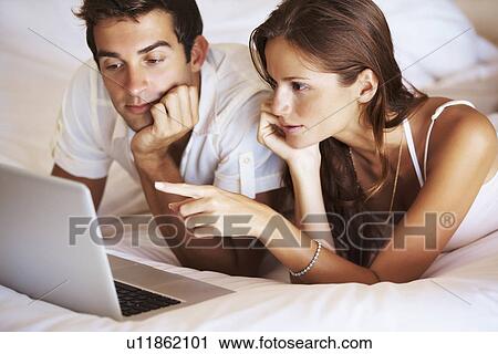 Couple using laptop while lying in bed View Large Photo Image Stock Image - Couple using laptop while lying in bed. Fotosearch
