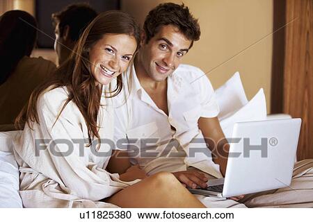 Stock Image - Couple using laptop while sitting on bed. Fotosearch