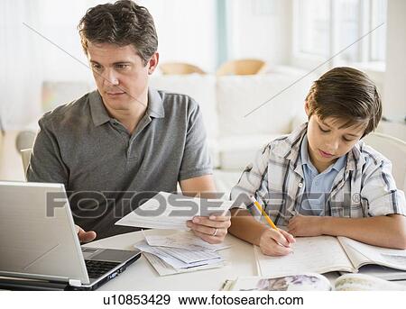 Stock Photo - Father working on laptop and son (8-9) doing homework. Fotosearch