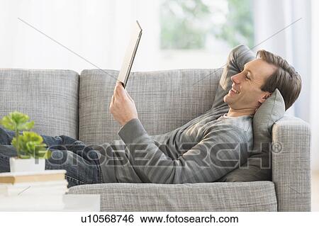 Man lying on sofa watching tablet pc View Large Photo Image Stock Photograph - Man lying on sofa watching tablet pc. Fotosearch
