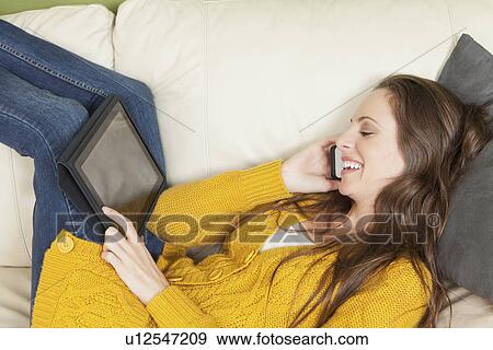 Woman lying on sofa with digital tablet and talking via cell phone View Large Photo Image Stock Photo - Woman lying on sofa with digital tablet and talking via cell phone. Fotosearch