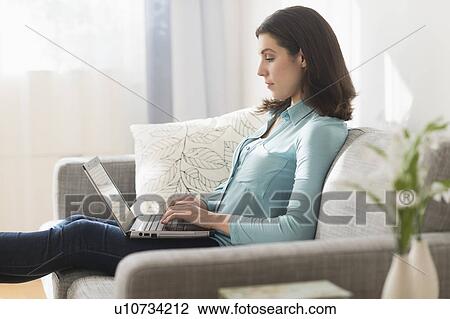 Stock Image - Woman sitting on sofa and using laptop. Fotosearch