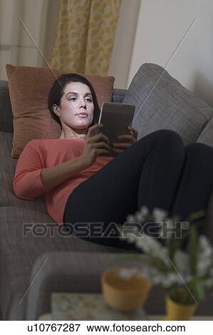 Stock Photo - Woman using digital tablet while lying on sofa. Fotosearch