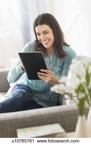 Stock Image - Woman using digital tablet while sitting on sofa. Fotosearch