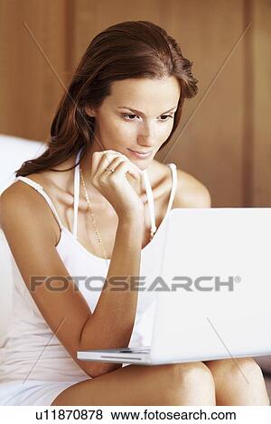 Stock Photo - Woman using laptop while sitting on bed. Fotosearch