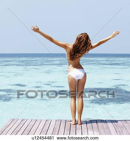 Stock Image - Young woman in bikini standing on jetty. Fotosearch
