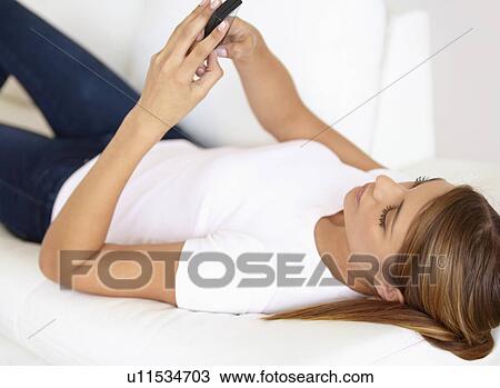 Stock Image - Young woman lying on bed and using cell phone. Fotosearch