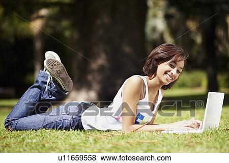 Stock Photo - Young woman lying on lawn in park and using laptop. Fotosearch