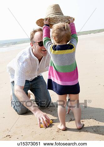 Stock Image - Girl putting hat on father at beach. Fotosearch